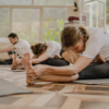 A side-view of a yoga class in a bright studio with large windows and a patterned wooden floor. In the foreground, a woman with blonde hair in a braid is performing a seated forward fold with one leg tucked in, holding her foot for a deep stretch. Behind her, two other participants are in similar seated stretching poses on their mats.