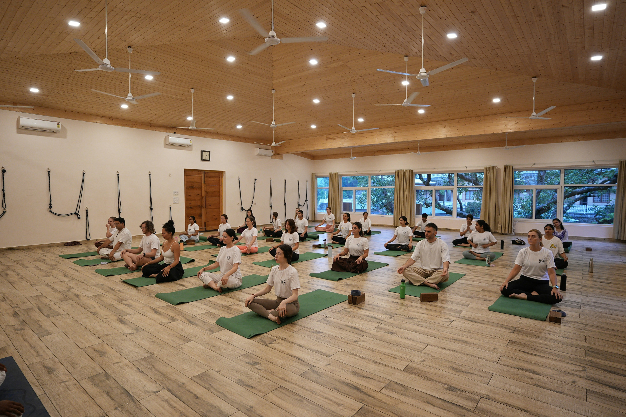 A diverse group of people sitting cross-legged in a meditative pose on green yoga mats. They are in a large, bright studio with light wood floors and a matching wooden ceiling equipped with several white ceiling fans and recessed lighting. Large windows in the background reveal greenery outside.