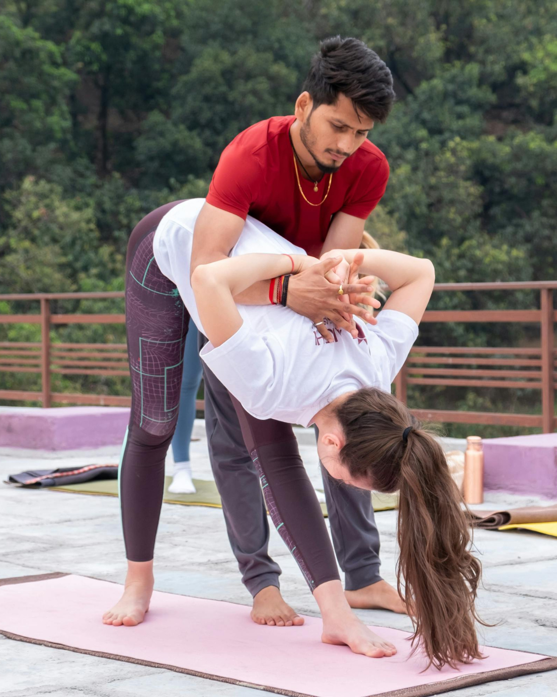 Yoga instructor assisting a woman in a deep forward bend/backbend stretch on a mat outdoors, helping improve flexibility and proper posture.