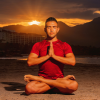 A man sitting in a cross-legged lotus yoga pose on a flat outdoor surface. He is wearing a red short-sleeved athletic shirt and has his hands in a prayer position at his chest with his eyes closed. Behind him, a vibrant orange and yellow sunset glows over the dark silhouette of a mountain range. The warm golden hour light illuminates the front of his body, casting a soft shadow behind him.
