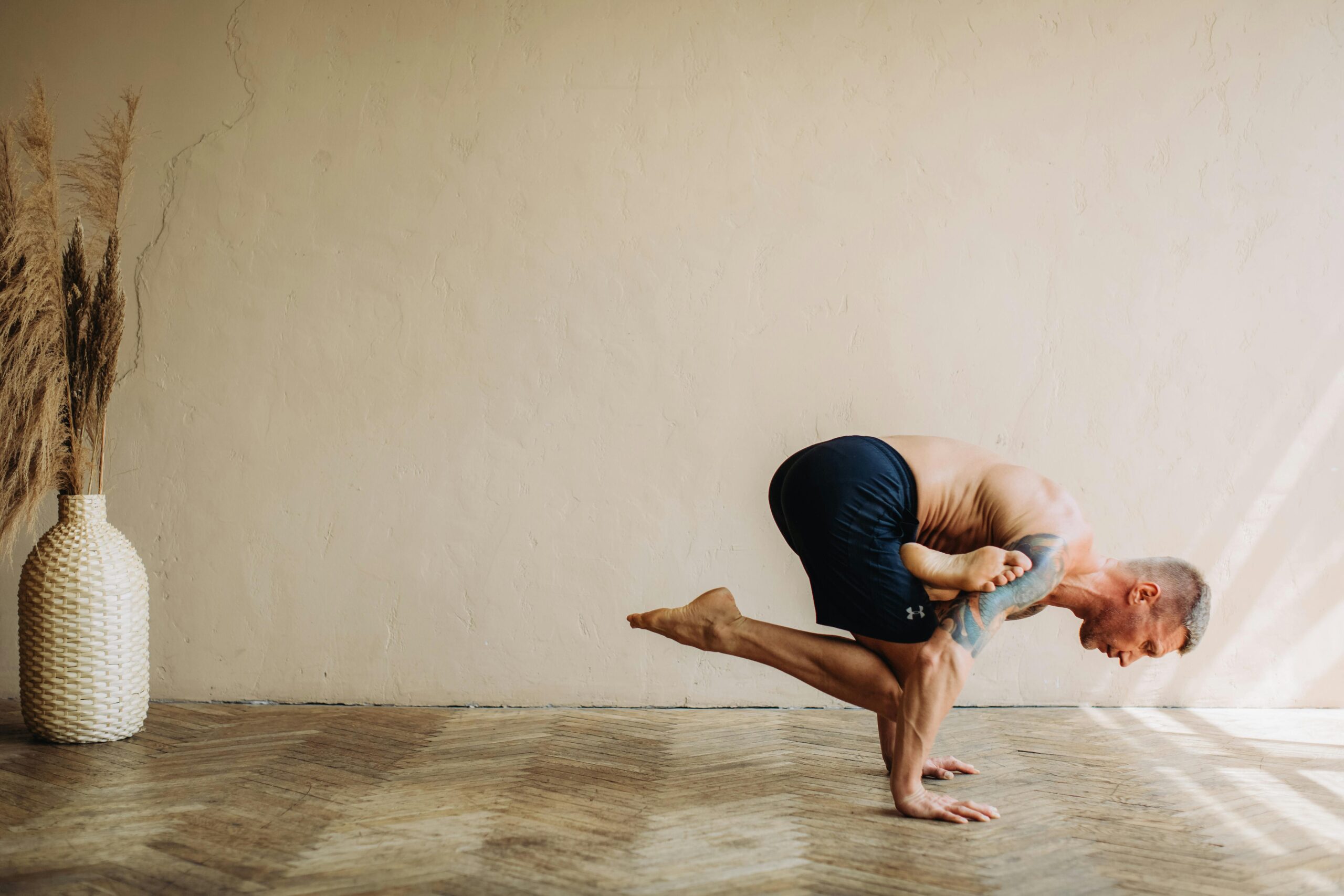 A side-profile view of a man with a tattooed arm performing an advanced arm balance yoga pose, a variation of Grasshopper or Dragonfly pose, in a minimalist studio. He is balanced on his hands with one leg tucked and the other extended behind him. The room features textured beige walls, a herringbone-patterned wooden floor, and a tall, woven vase with dried pampas grass.
